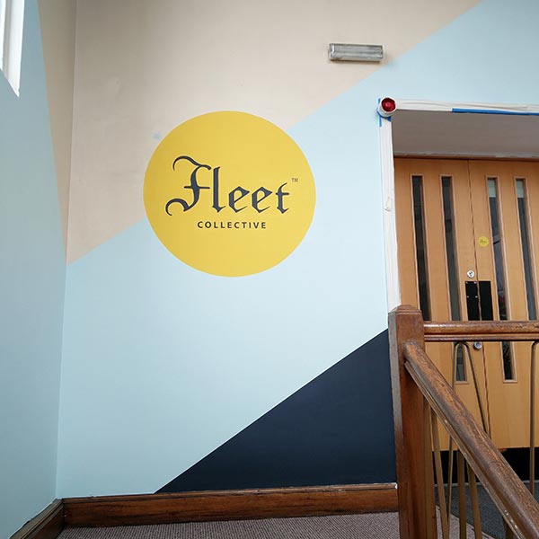 Yellow circle Fleet Collective logo sits over light blue and dark blue stripes on a wall.