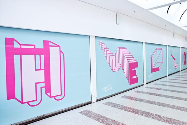 Woman with trolley bag walks past huge 'hello' in giant letters in a shopping centre corridor.