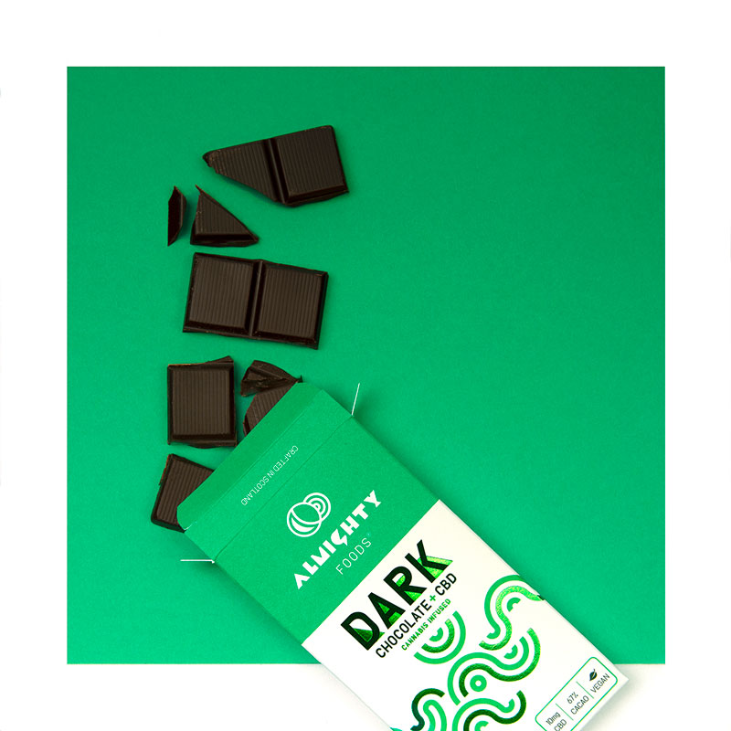 White and green chocolate box for the dark chocolate with CBD. The box is sitting on a green background with chocolate pieces spilling out of the top.