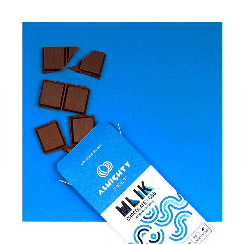 Blue and white chocolate box for the mlik chocolate with CBD. The box is sitting on a blue background with chocolate pieces spilling out of the top.