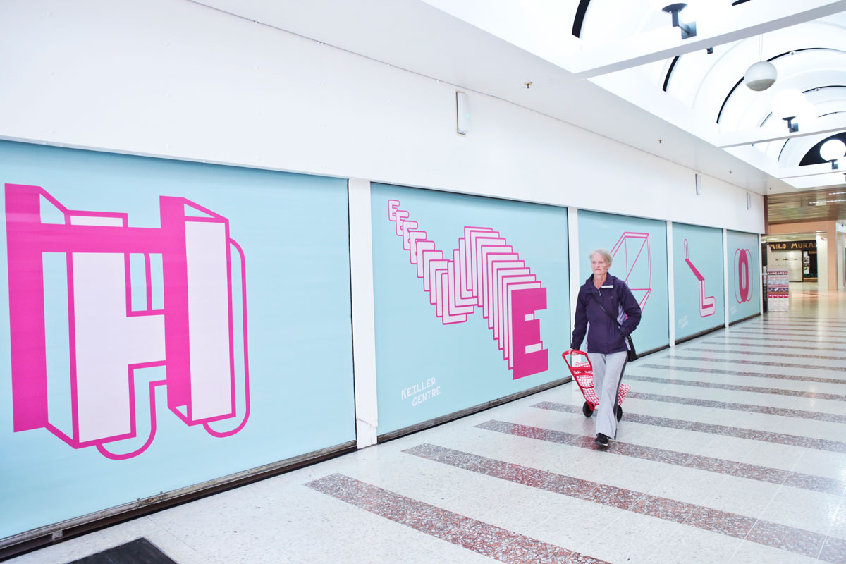 Woman with trolley bag walks past huge 'hello' in giant letters in a shopping centre corridor.