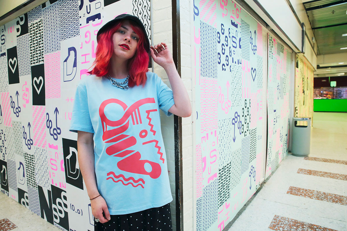 Woamn with bright red hair holds a bucket hat on her head while modeling a graphic t-shirt. Set on the backdrop of typographics pasted up posters with a shopping centre.