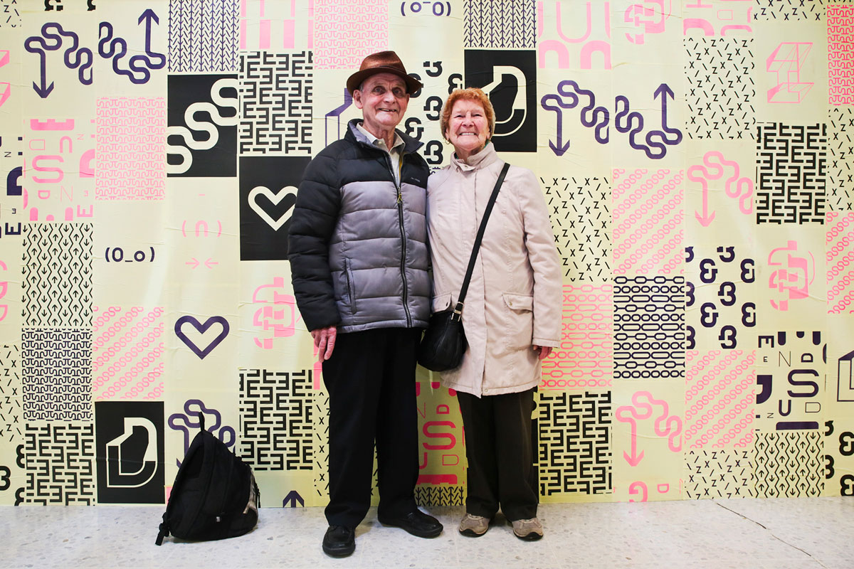 Smiling eldery couple stand in front of a wall of typographic posters.
