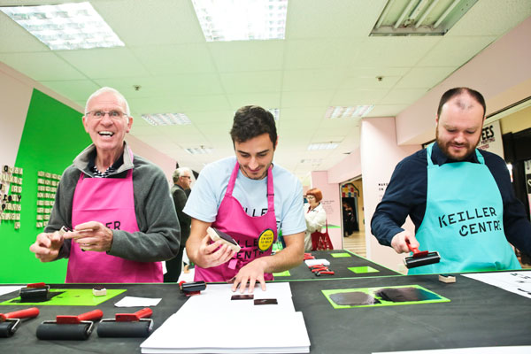 3 men side by side in aprons inking up and printing with lettering blocks