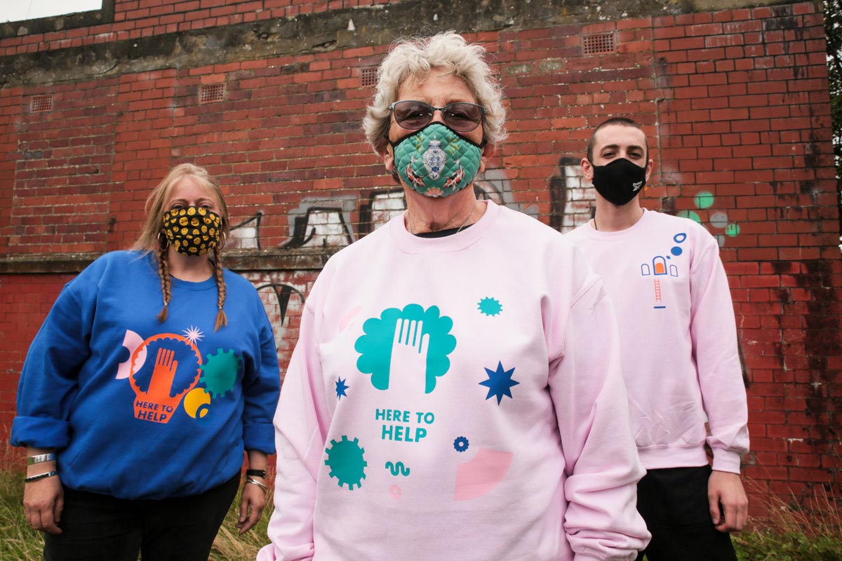 3 volunteers wearing masks show of their self-designed sweatshirts with the words here to help.